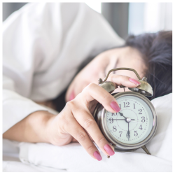 Vrouw liggend in bed, met een wekker in haar handen en roze nagels, net klaar met haar nachtelijke huidverzorgingsroutine