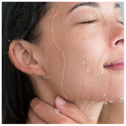 Jonge vrouw met een natte huid die onder de douche staat, terwijl water over haar gezicht stroomt